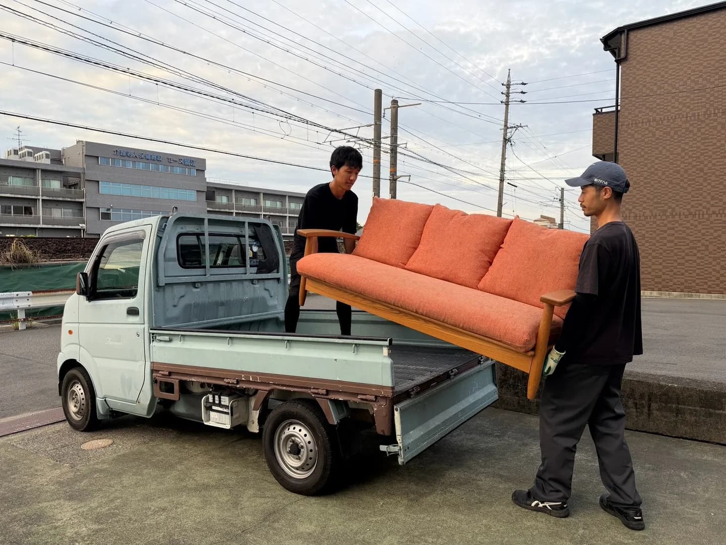 オレンジ色の木枠ソファを軽トラックの荷台付近で2人のスタッフが支える様子